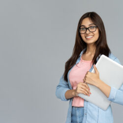 Smiling indian young woman college university school student wear glasses holding laptop computer isolated on grey studio background, happy teen girl looking at camera stand with device, portrait