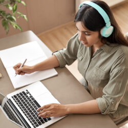 Indian girl student wear headphones learning online watching webinar class looking at laptop computer elearning remote lesson making notes or video calling virtual conference meeting teacher at home.