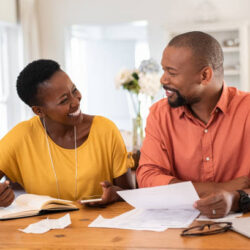 Cheerful mature couple sitting and managing expenses at home. Happy african man and woman paying bills together and managing budget. Black smiling couple checking accountancy and bills while looking at each other.